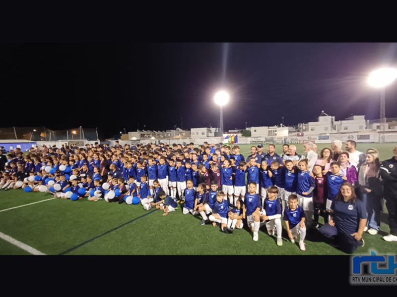 Equipo de fútbol infantil y adultos posando juntos en un campo de fútbol iluminado por luces de foco nocturnas.