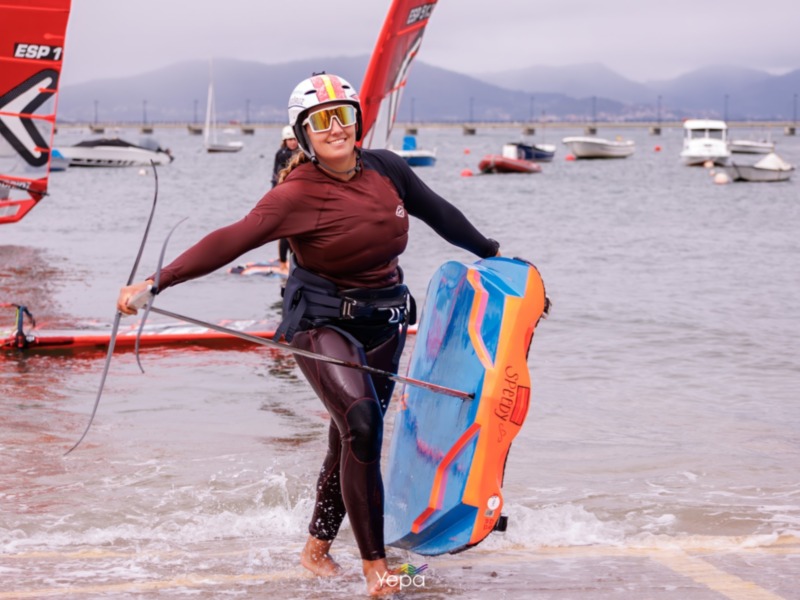 Una deportista de windsurf sonríe mientras se prepara para el salto, con el mar y las montañas en el fondo.