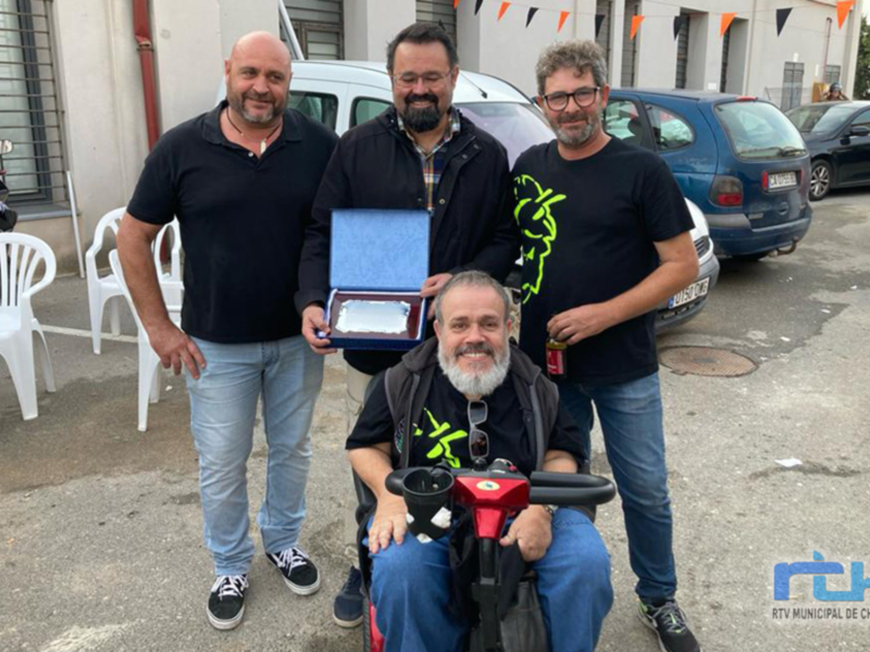 Una persona en silla de ruedas sostiene un triciclo, rodeada por tres hombres que le entregan una medalla. En el fondo, se observa un edificio con banderas y sillas blancas al aire libre.