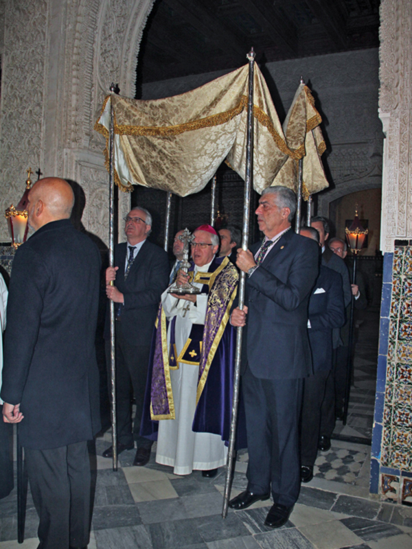 Imagen de un evento formal en una iglesia o palacio, con varios hombres vestidos formalmente. Algunos llevan antorchas y otros están en trajes oscuros, uno con un hábito religioso. La decoración es elegante y el ambiente parece ser de una celebración o ceremonia importante.