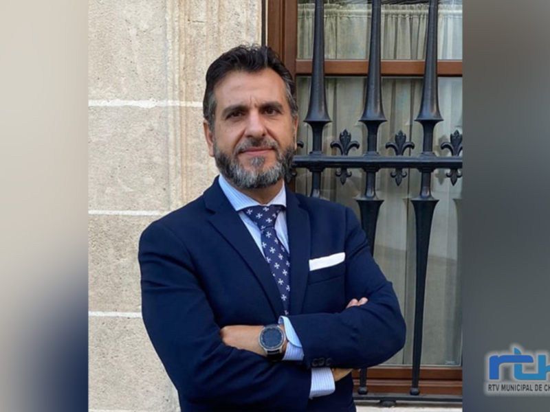 Retrato de hombre en traje de negocios con brazos cruzados, frente a una ventana con rejas, en un entorno urbano.