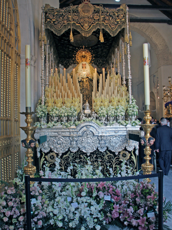 Procesión religiosa con imagen de Virgen María en un paso ornado con flores y candelabros, en un entorno eclesiástico.