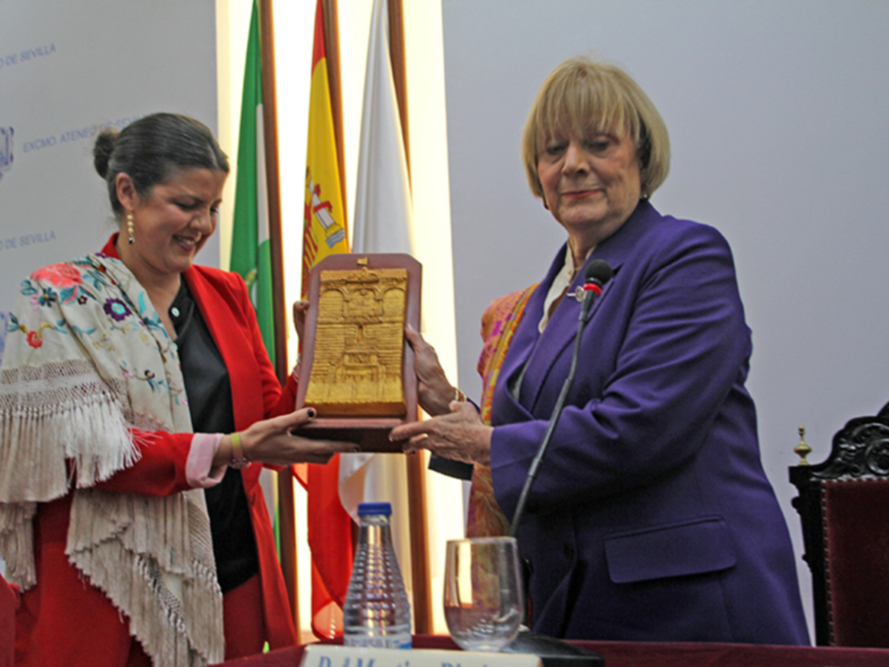 Mujeres intercambian un trofeo en un evento formal con banderas de España y Andalucía en el fondo.