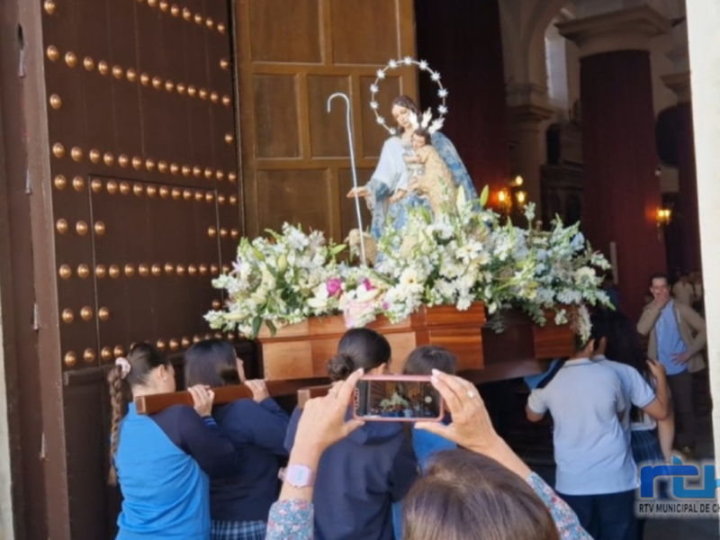 Fotografía de una procesión religiosa en una iglesia, con una imagen de la Virgen María y el Niño Jesús en el centro, rodeada de flores y seguida por fieles que la fotografían.