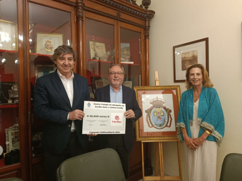 Señor y señora sonríen mientras sostienen un certificado blanco con el texto "B 36.000 euros" y una imagen de un escudo real en el fondo. La mujer está vestida con un vestido blanco y una chaqueta azul, mientras que el hombre lleva una camisa blanca y un traje negro. La imagen se encuentra en un entorno formal con una vitrina de madera y objetos decorativos.