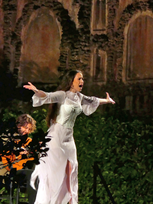 Cantante en escenario con vestido blanco, interpretando con pasión en un entorno histórico.