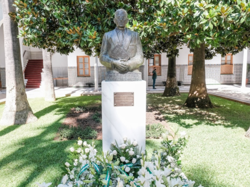 Estatua de un hombre en un jardín con flores blancas y árboles grandes. El jardín está rodeado por una estructura con columnas y una escalera en el fondo.