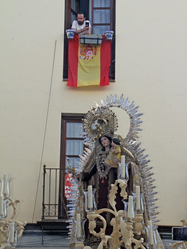 Procesión con estandarte español y imagen de la Virgen del Carmen en un entorno urbano.
