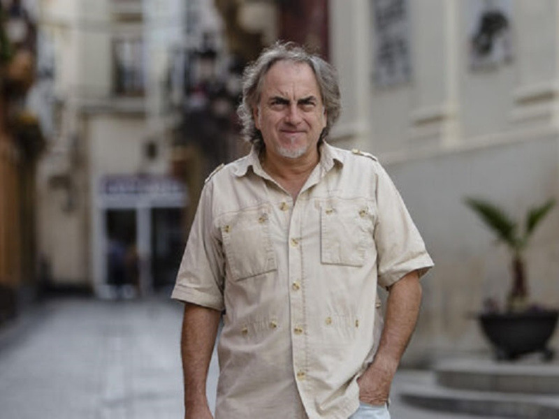 Un hombre con una camisa beige posa frente a una fachada de edificio con una planta en el fondo.
