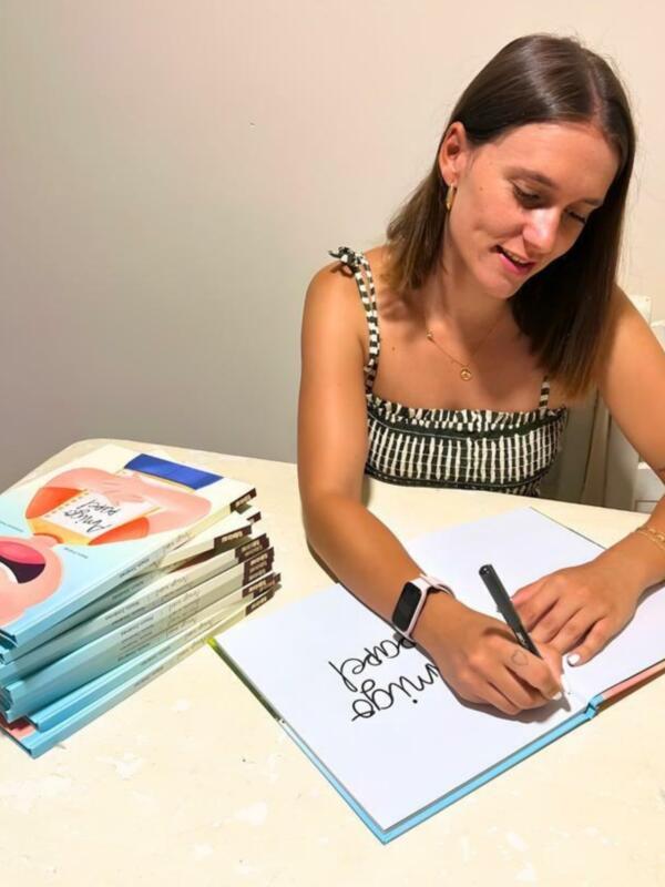 Una mujer escribe en un cuaderno mientras está sentada en una mesa con una pila de libros.
