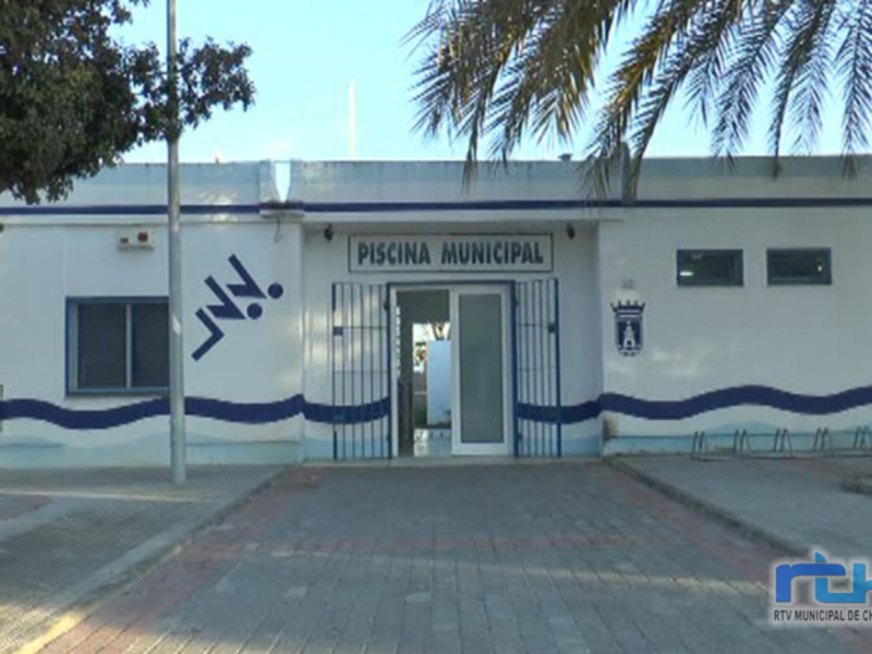 Piscina Municipal ubicada en la ciudad de Chipiona, España. Edificio blanco con azulejos azules y una palmera en el frente.