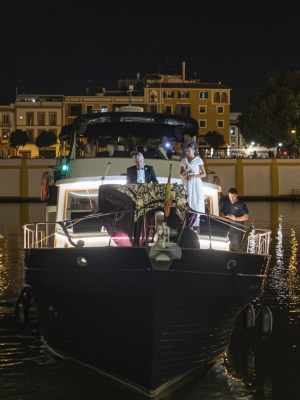 Una velada nocturna en barco con luces y figuras en el agua, frente a edificios iluminados.