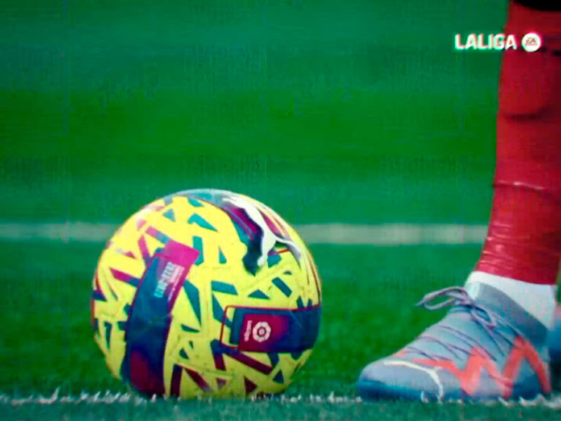 Detalle de un balón de fútbol en el césped de un campo de LaLiga, con una parte del pie del jugador en primer plano.