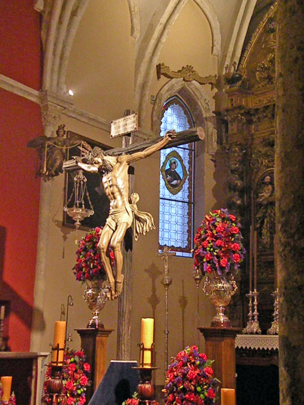 Se muestra una iglesia con un crucifijo en el centro, rodeado de flores y velas. La arquitectura es gótica con detalles dorados y vitrales. En el fondo, se puede ver una ventana con un diseño de ángeles y un altar decorado.