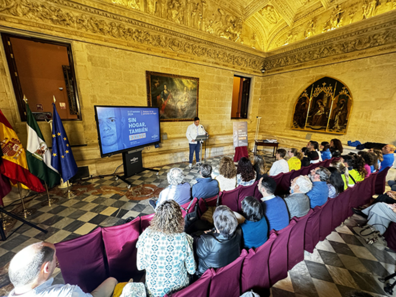 A un grupo de personas sentadas en filas, escuchando a un hombre que está hablando frente a una pantalla con texto. La sala tiene decoración clásica, con estatuas y murales en el techo. En la pared hay banderas de España, México y la Unión Europea. La atmósfera parece ser formal o educativa.