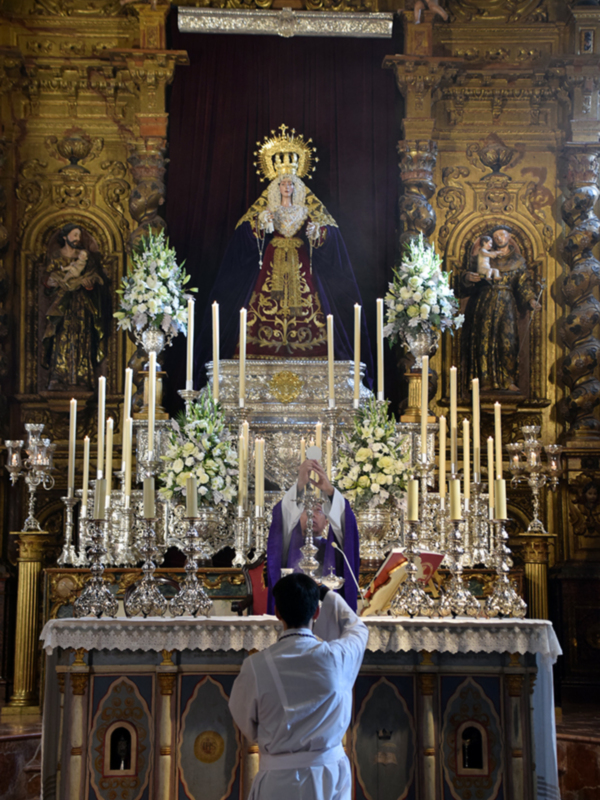 Sacerdote oficiando una misa en un altar ornado con flores y candelabros, en un templo con detalles barrocos.