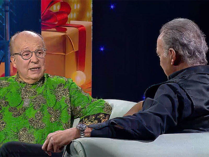 Entrevista en estudio con dos invitados sentados en sillones blancos, uno con camisa verde brillante y otro en chaqueta negra, en un ambiente festivo con luces y regalos de fondo.