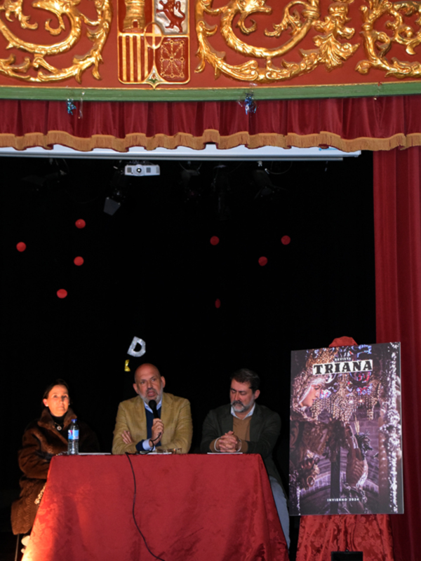 Tres personas sentadas en una mesa roja con un escudo en el fondo, posiblemente en un evento formal o ceremonial.