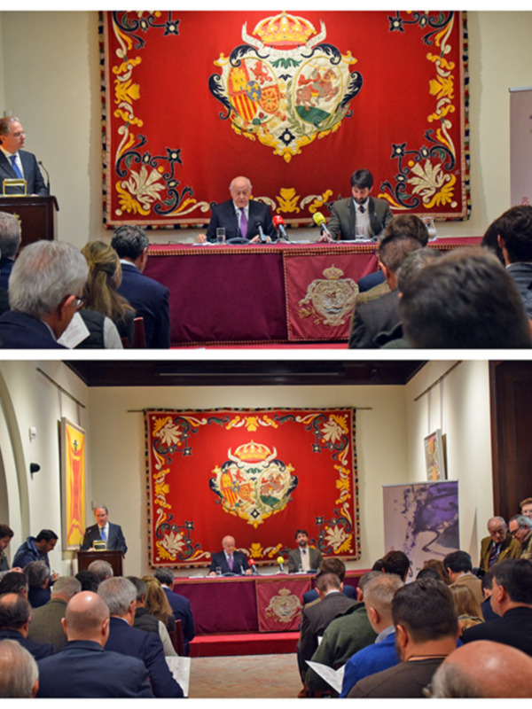 Conferencia de prensa en un salón con escudo real español, con participantes en la mesa y público asistiendo.