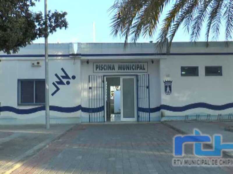 Entrada de la Piscina Municipal de Chipiona, con su fachada blanca y azul, rodeada de palmeras y un entorno urbano.