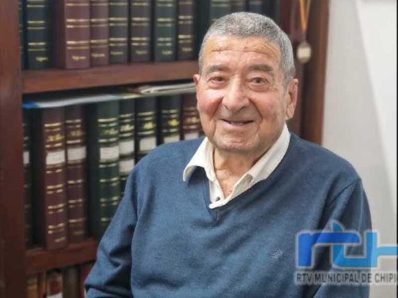 Un hombre mayor sonriendo frente a una estantería llena de libros. La imagen está acompañada por el logotipo de RTVMUNICIPALIDAD DE CHIPIONA.