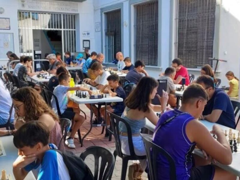 Jugadores de ajedrez en una competición al aire libre en un patio de la escuela, con varias mesas y sillas, y edificios en el fondo.