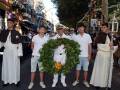Un grupo de personas en una procesión, incluyendo un hombre con un sombrero militar y un niño sosteniendo una corona de laurel, caminan por una calle con edificios y árboles a lo largo.