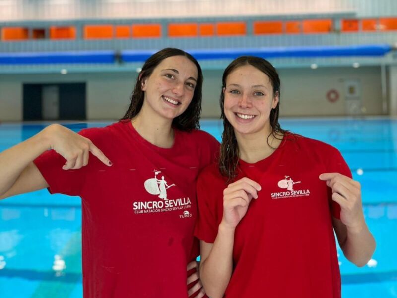 Dos jóvenes posan en una piscina, sonrientes y con camisetas rojas de Sincro Sevilla. La imagen se centra en ellos frente a una piscina interior con azulejos verdes y naranjas.