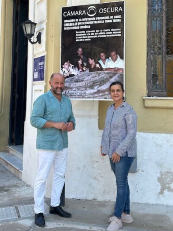 Dos personas posan frente a una pared amarilla con un cartel de la Cámara Oscura. La imagen parece ser parte de una exposición o evento cultural.