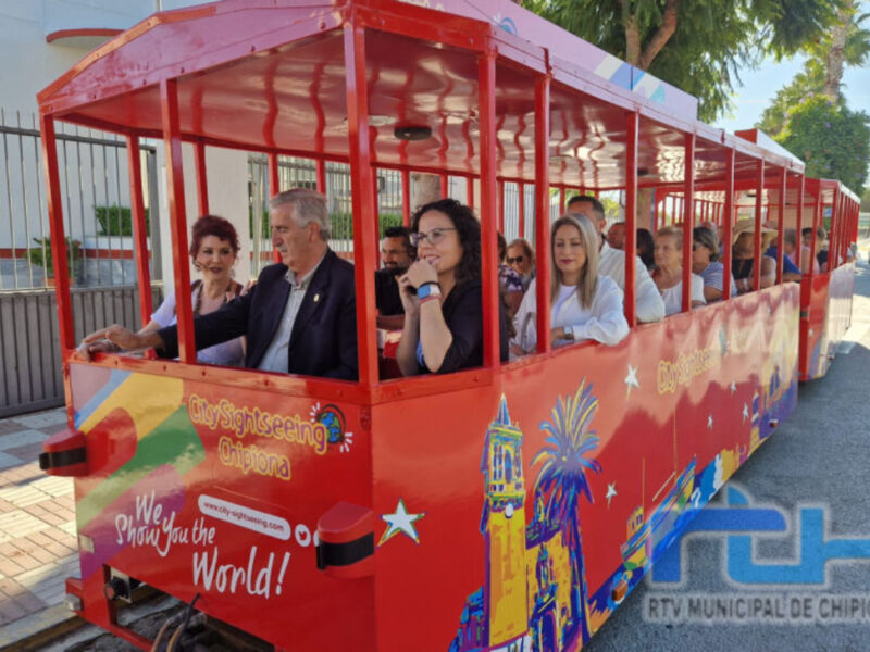 Un tren turístico rojo con dibujos de edificios y estrellas, lleno de personas. La imagen muestra una ruta turística en Chipiona, con el lema "Smile the World" y la marca de RTV Municipal de Chipiona.