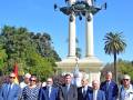 Una imagen de una fila de personas vestidas con trajes formales, posando frente a un monumento alto y elegante. El cielo es azul claro y hay árboles verdes al fondo, lo que sugiere un entorno tranquilo y formal.