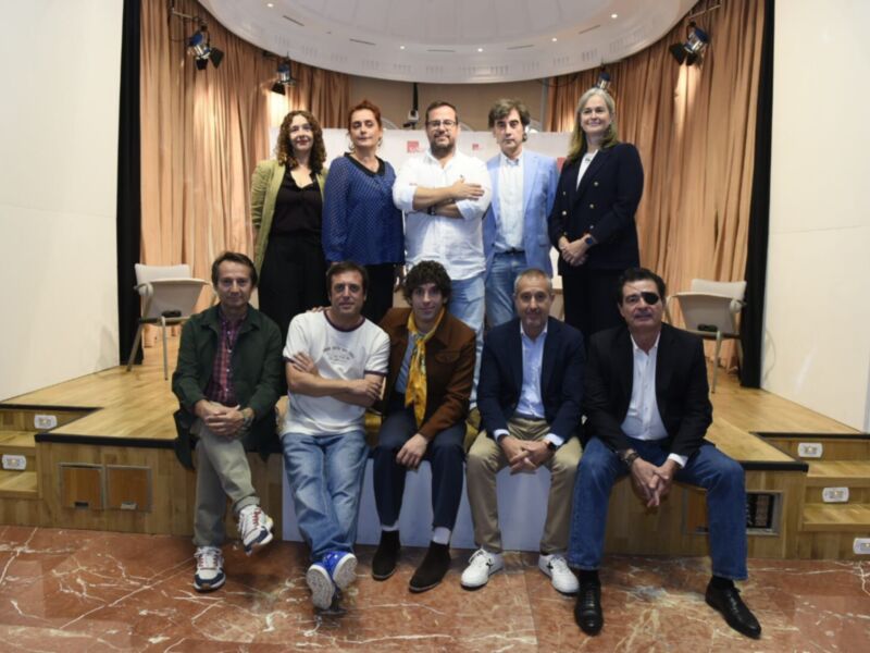 Un grupo de personas posa en una sala con cortinas y un escenario. La imagen muestra a siete individuos, cuatro sentados en las escaleras y tres de pie detrás. La sala tiene una decoración formal con un suelo de mármol y una estructura en el fondo.