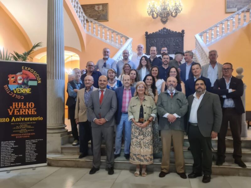 Grupo de personas posando frente a un póster promocional para la exposición "Julio Verne". La imagen se tomará en un entorno con columnas y una escalera, iluminada por una lámpara de araña.
