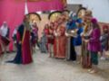 Una celebración festiva con personajes vestidos de trajes tradicionales, posiblemente inspirados en la época medieval. Los personajes están distribuidos alrededor de un área cubierta por una estructura roja y dorada, que parece ser parte de un escenario o decoración para la celebración. La escena se desarrolla en una calle adoquinada, con algunas personas observando la celebración desde el fondo.