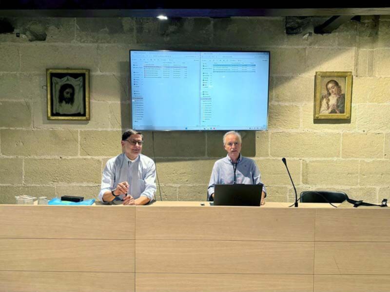 Dos hombres sentados en una mesa frente a un televisor proyectando código, con dos cuadros decorativos en la pared.