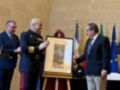 En una ceremonia formal, un oficial militar y otros dos individuos en trajes de negocios se encuentran alrededor de un stand con una imagen de un hombre en uniforme. La escena está ambientada en un entorno interior con una pared de ladrillo y banderas en el fondo.