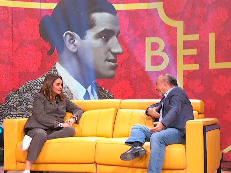 En un programa televisivo, dos personas se sientan en un sofá amarillo frente a una pantalla que muestra una imagen de un hombre con el nombre "BELM" en letras grandes. La escena transmite una conversación o entrevista formal, con un fondo decorado en rojo y dorado.