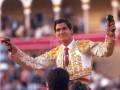 Un torero en traje tradicional, con un fondo de arcos dorados y espectadores en la arena.