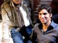Dos hombres sonrientes, uno con chaqueta beige y el otro en polo azul. Ambos están sentados en una escalera de cristal iluminada por luces doradas.