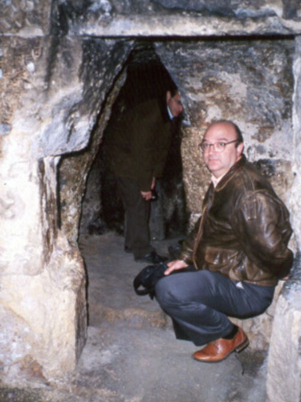 Dos hombres explorando un túnel de piedra, uno con chaqueta marrón y gafas, el otro con camisa negra.