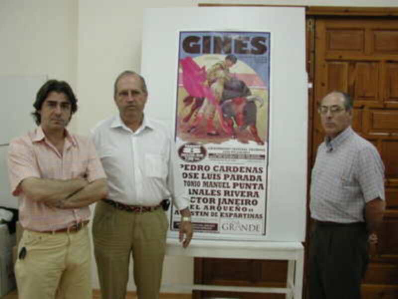 Tres hombres posan junto a un cartel de toros con el nombre 'Gines' y detalles de la corrida.