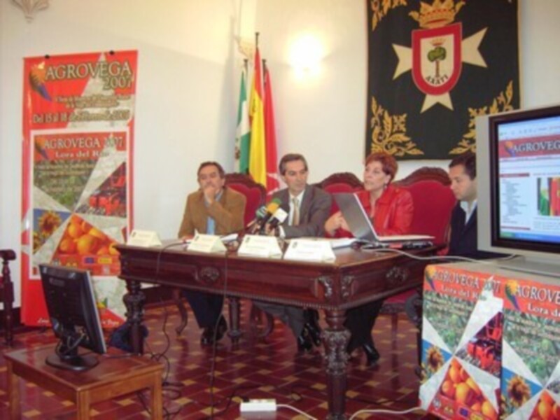 Una reunión formal en una sala con una mesa central, dos personas sentadas en sillas rojas y otra en una silla de madera. Hay carteles informativos y banderas en el fondo. El ambiente es formal y profesional.