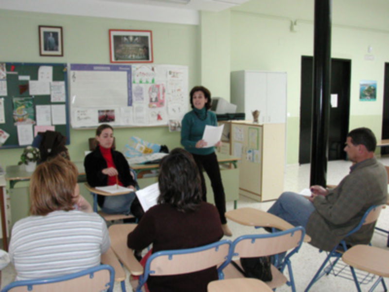 Clase en un aula con estudiantes sentados y una profesora hablando. La pared está decorada con carteles y un calendario.