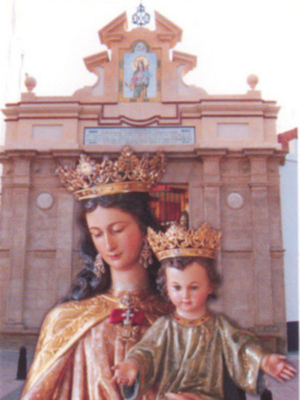 Estatua religiosa de una mujer con un niño en sus brazos, ambos vestidos con trajes dorados y coronas. El fondo muestra un arco con inscripciones y una imagen en el centro.