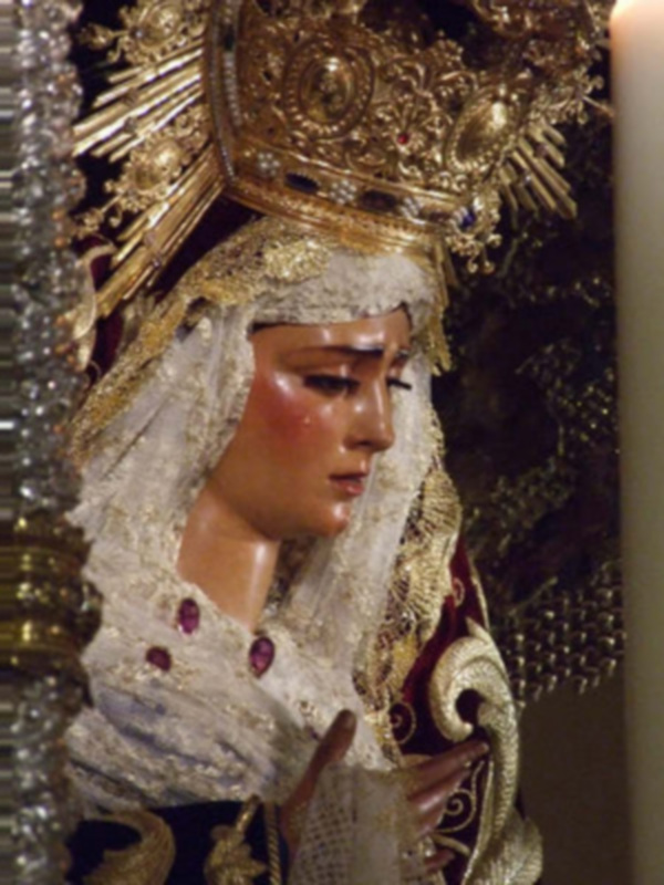 Una figura religiosa vestida con trajes elaborados, incluyendo un tocado dorado y una capa de color rojo. La figura parece estar en un entorno ceremonial o religioso, posiblemente durante una procesión.