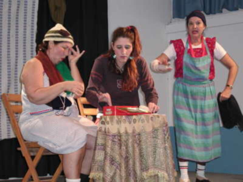 Tres personas en escena, una con bufanda y vestido blanco, otra con cabello rojo y vestido rojo, y una tercera con vestido a rayas. La escena parece ser de un teatro o una representación, con una silla y una caja en el centro.