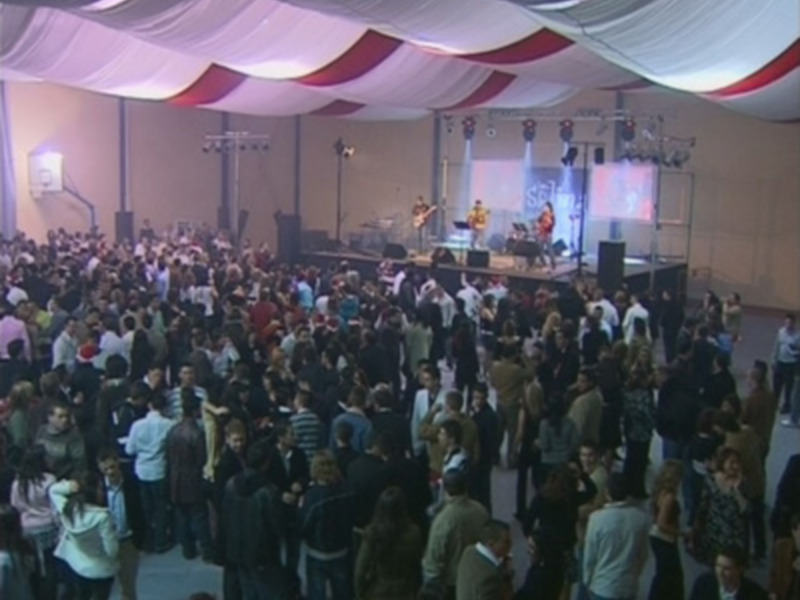Una multitud en una fiesta o evento, con un escenario iluminado y decoración de colores rojo y blanco en el techo.