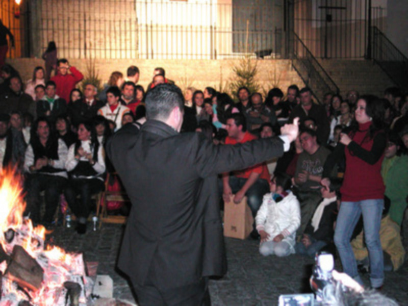 Una multitud asiste a una celebración nocturna alrededor de un fuego, con personas vestidas casualmente y una figura en el centro dirigiendo la reunión.
