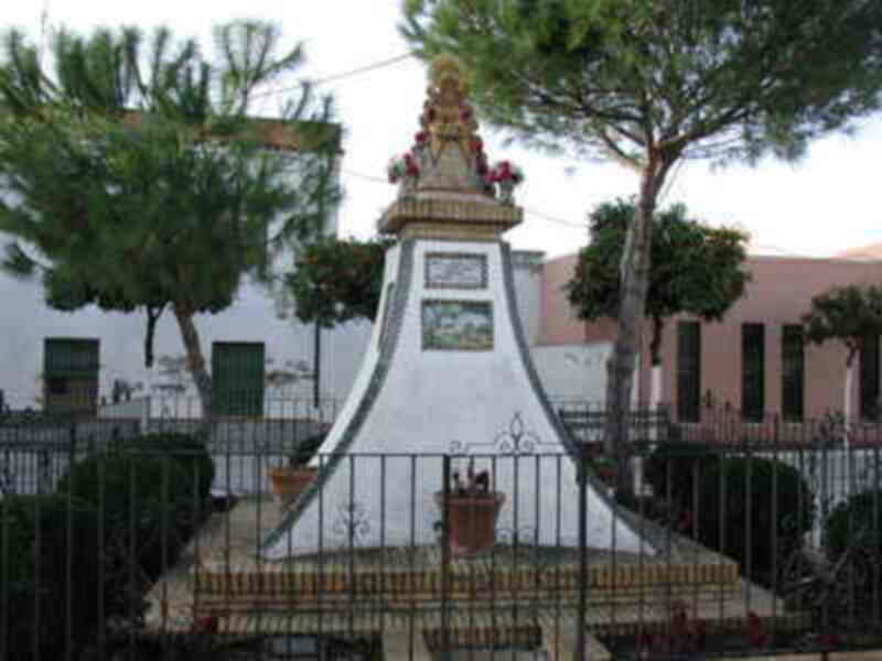 Monumento con flores y un escudo en el centro, frente a una casa blanca.