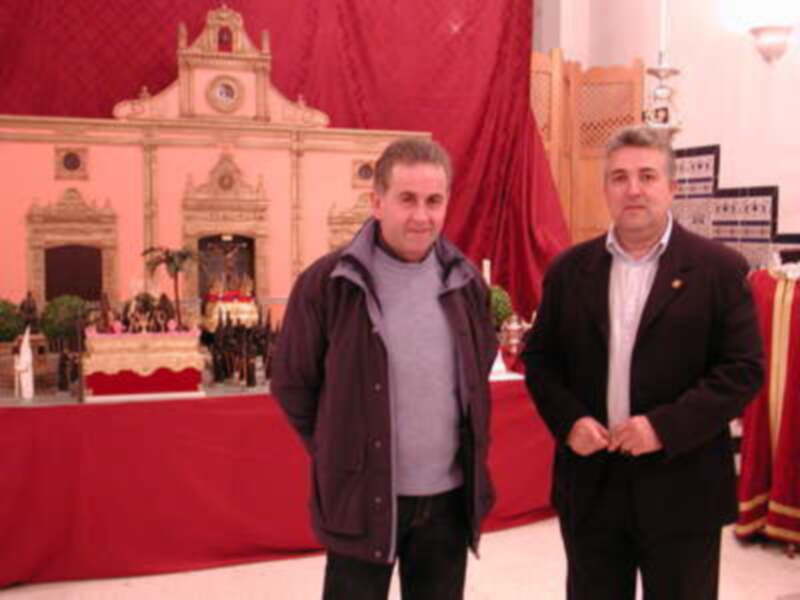 Dos hombres posan en frente de una iglesia con decoración roja y dorada. La imagen muestra un ambiente festivo, probablemente durante una celebración religiosa o social.